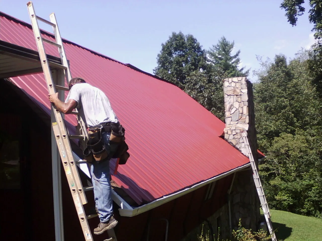 Roofer on a ladder beside a red metal roof and stone chimney — Soffit & Fascia Repair by Roofing Brothers in Blodgett